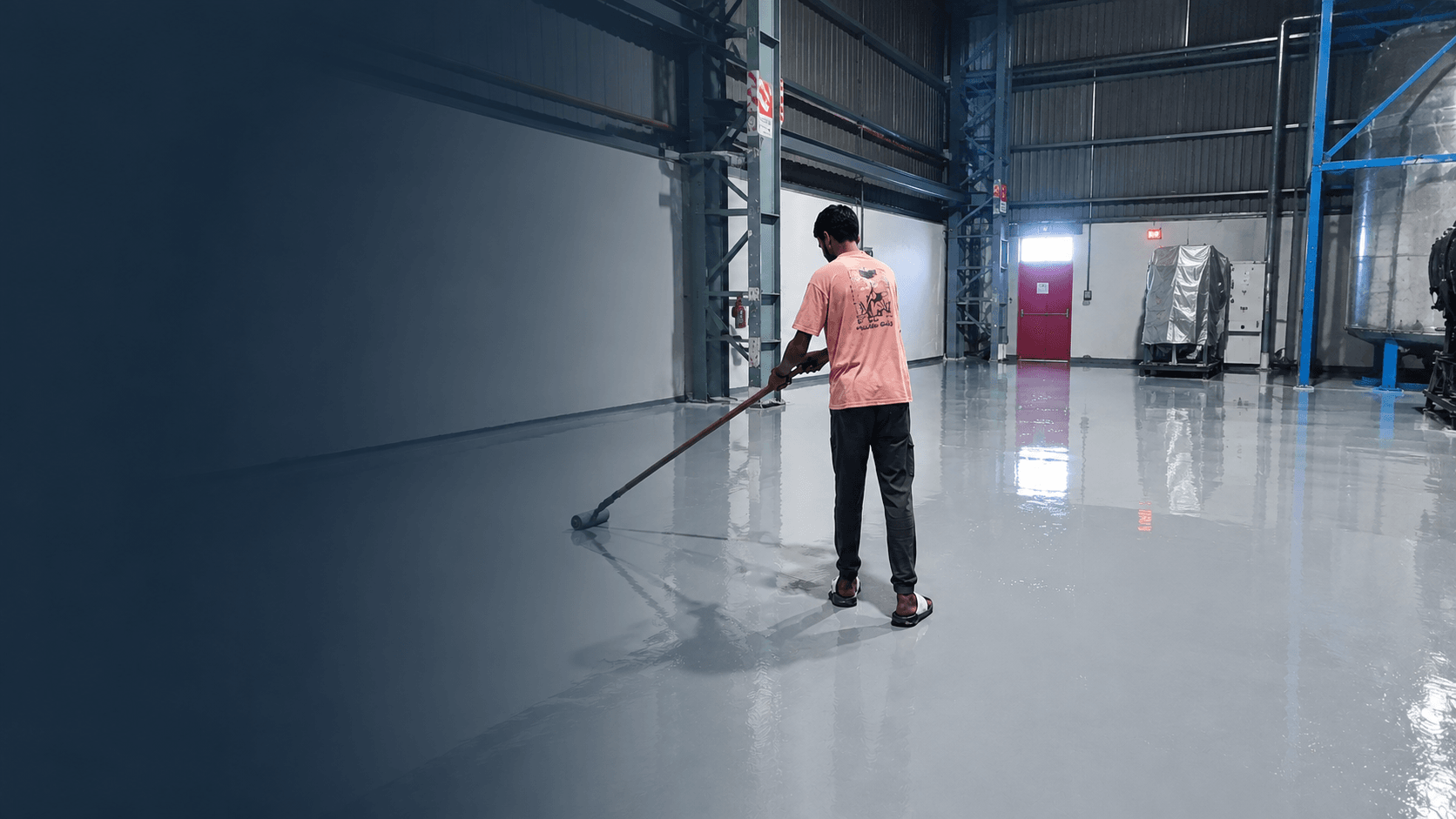 An applicator spreading epoxy floor coating with a squeegee across the floor of an industrial plant, the finished surface reflecting the roof structure behind him.