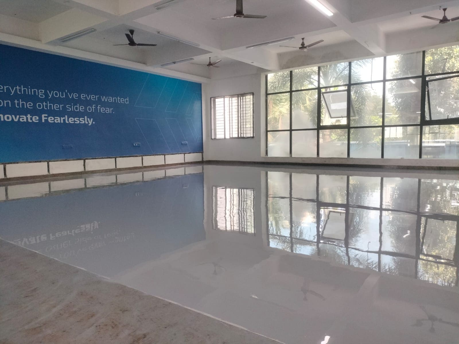 A finished grey epoxy floor in a bright office hall with a large window wall and a blue branded feature wall.